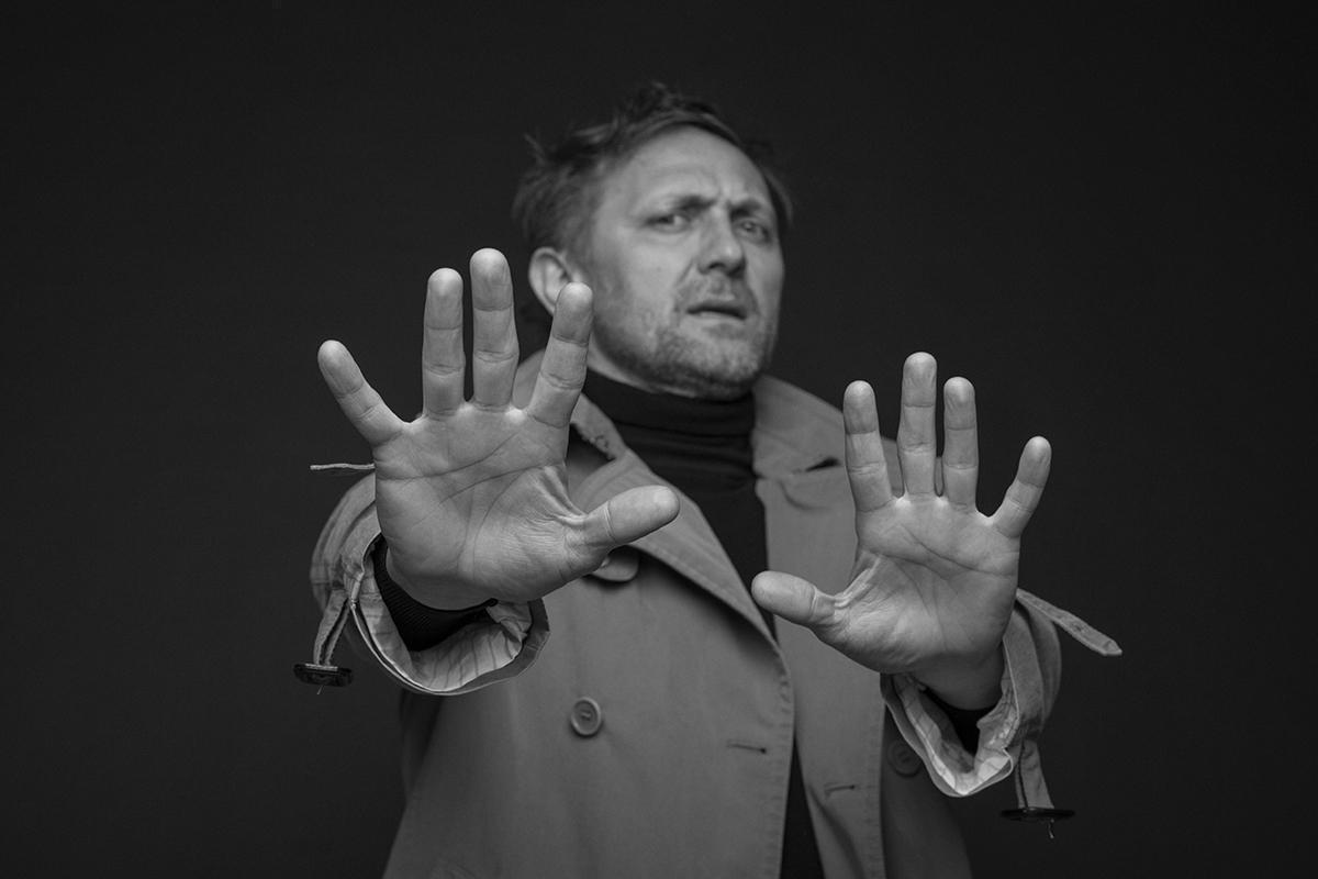 Andrzej Chyra w płaszczu, trzymający ręce w stronę obiektywu.