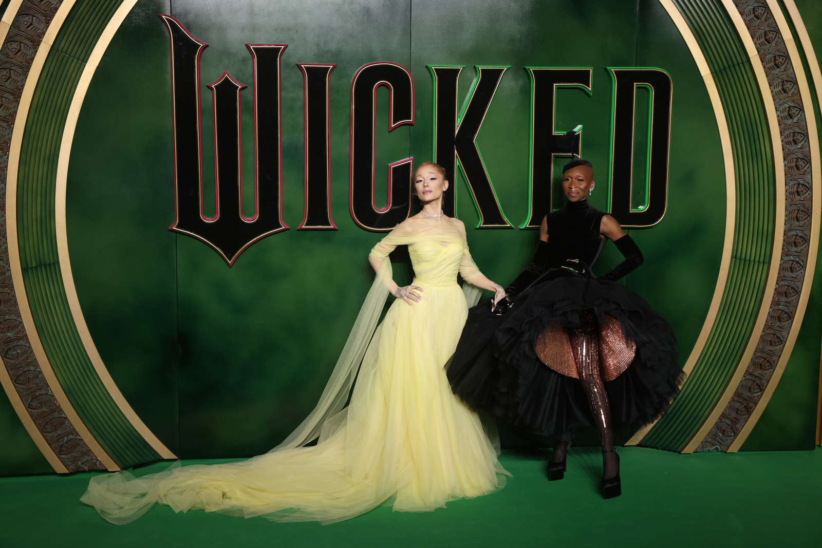 Ariana Grande i Cynthia Erivo pozujące na tle zielonego logo „Wicked", jedna w żółtej sukni, druga w czarnej, eleganckiej kreacji.
