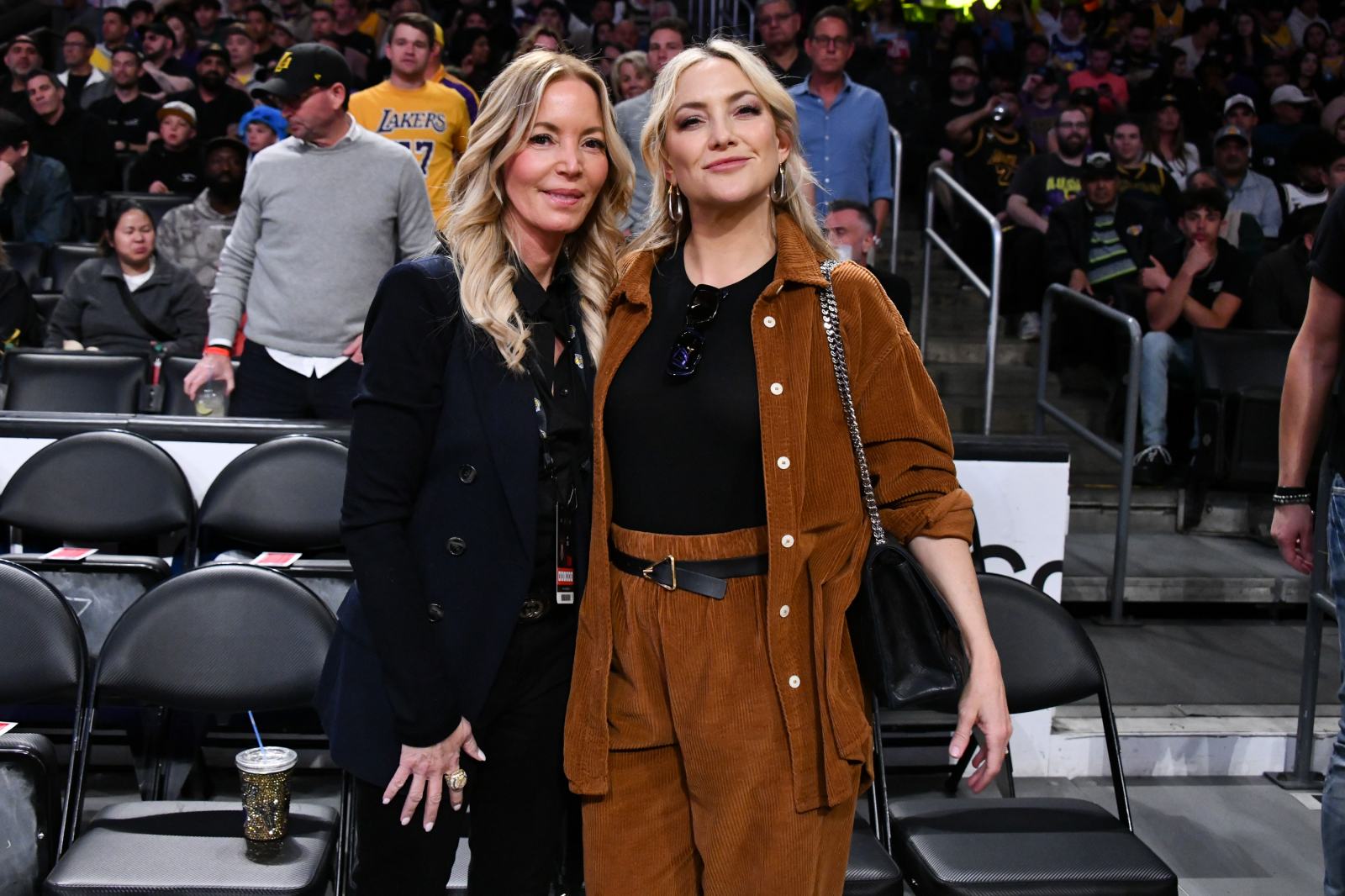 Kate Hudson pozująca z przyjaciółką na meczu, w stylowych strojach, w tle tłum kibiców.