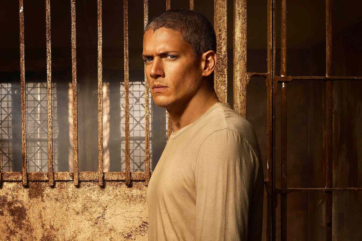 Wentworth Miller, wcielający się w rolę Michaela Scofielda, stoi przy kratowej barierze, ma na sobie beżową koszulkę i patrzy poważnie w stronę kamery.