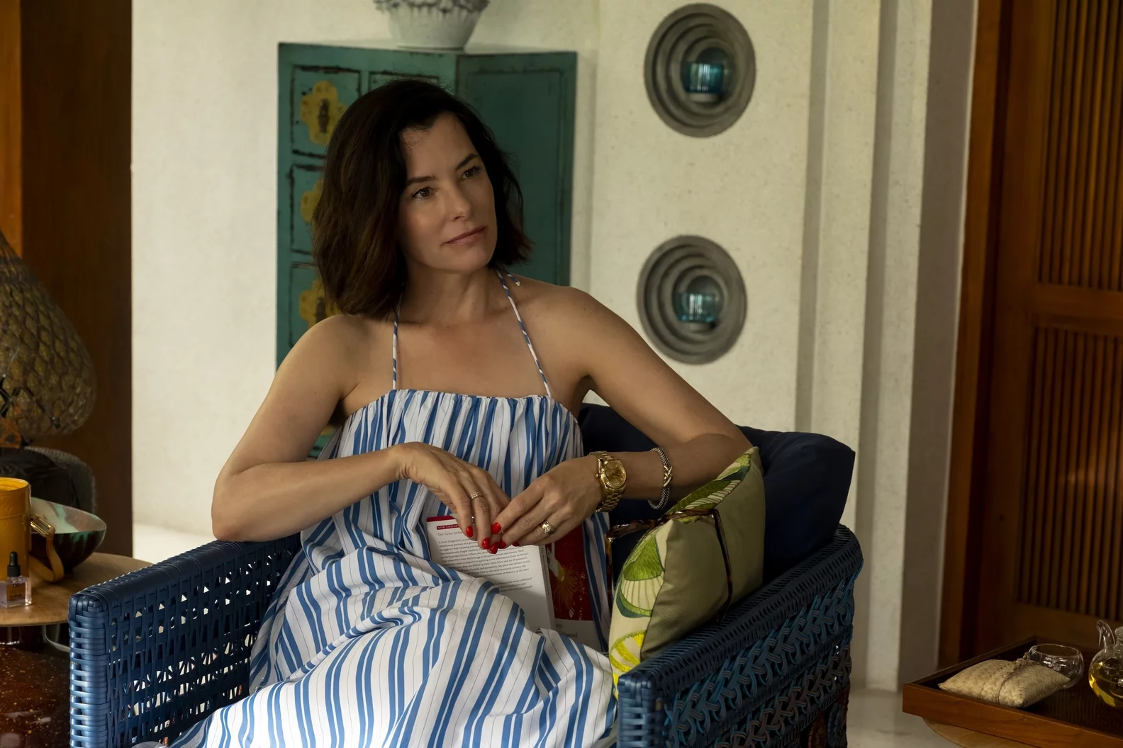Parker Posey jako Victoria w stylowej sukience siedzi na tarasie, w tle elementy dekoracyjne z serialu "Biały Lotos 3". Ma czerwone paznokcie.