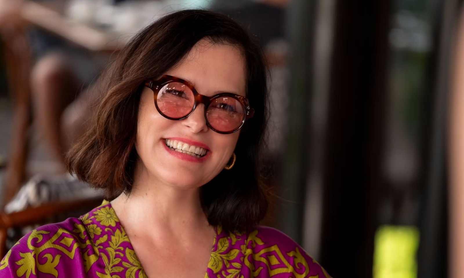 Parker Posey z uśmiechem, w okularach, w kolorowej sukience. Kadr z serialu biały lotos 3. Parker ma usta w odcieniu orchidei pomalowane pomadką Chanel