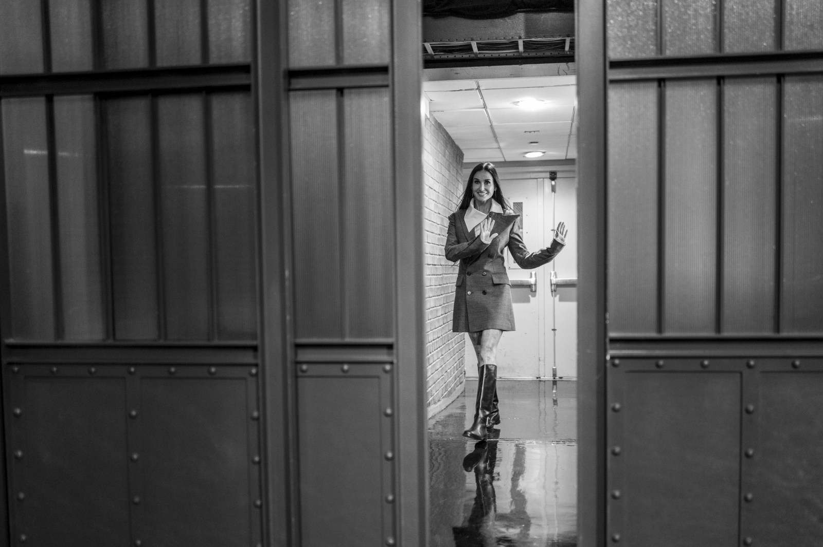 Demi Moore w eleganckiej marynarkowej sukience mini i oficerkach stoi w drzwiach, uśmiechając się i machając ręką. Scena w odcieniach szarości.