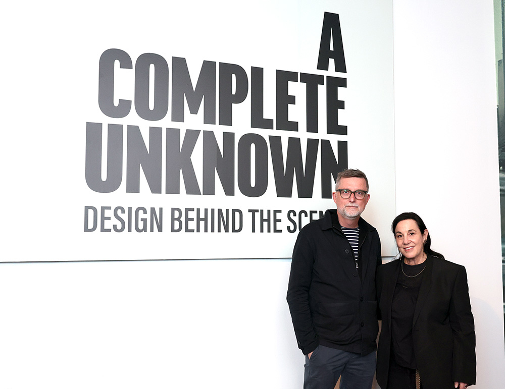 Dwoje ludzi stoi obok napisu "A COMPLETE UNKNOWN DESIGN BEHIND THE SCENES" w jasnym wnętrzu.