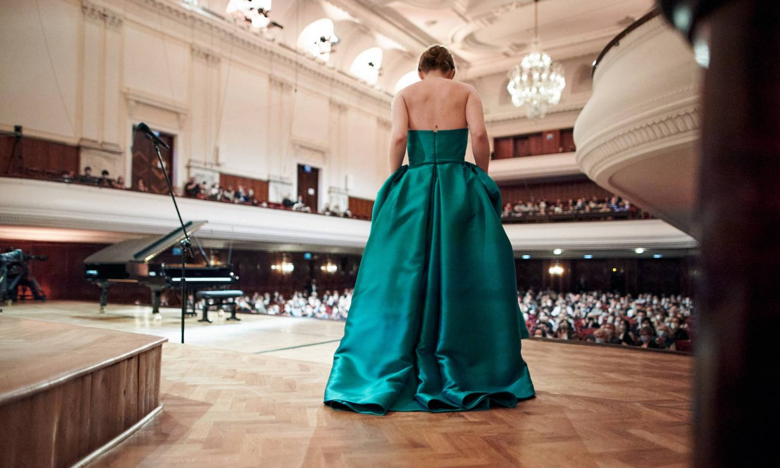 Kadr z filmu "Pianoforte", przedstawiający kobietę w turkusowej sukni stojącą na scenie przed publicznością, obok fortepian.
