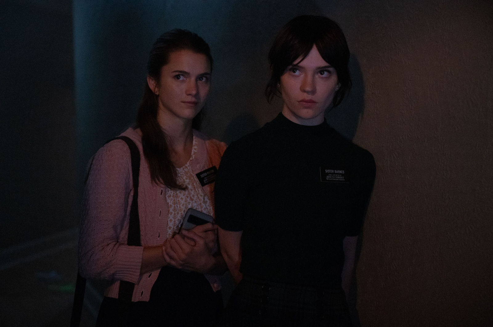 Chloe East i Sophie Thatcher w filmie „Heretic”, stoją obok siebie w mrocznym korytarzu, z poważnymi wyrazami twarzy.