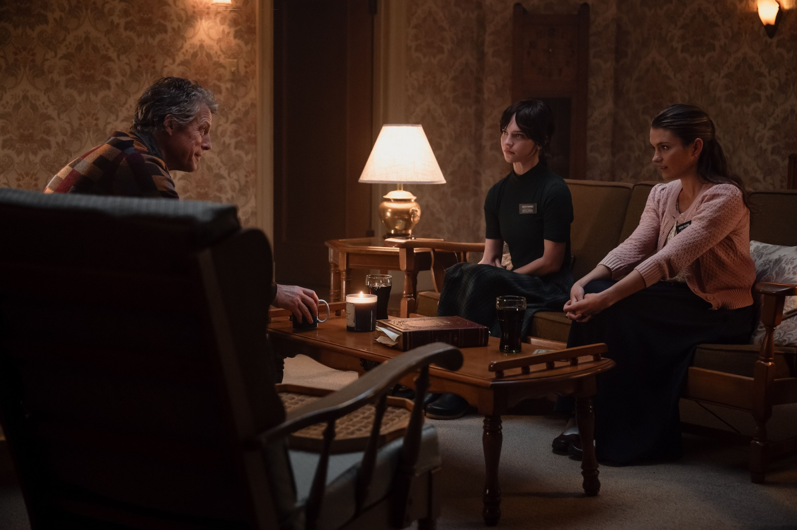 Hugh Grant, Chloe East i Sophie Thatcher w filmie „Heretic”, rozmawiają w przytulnym wnętrzu.