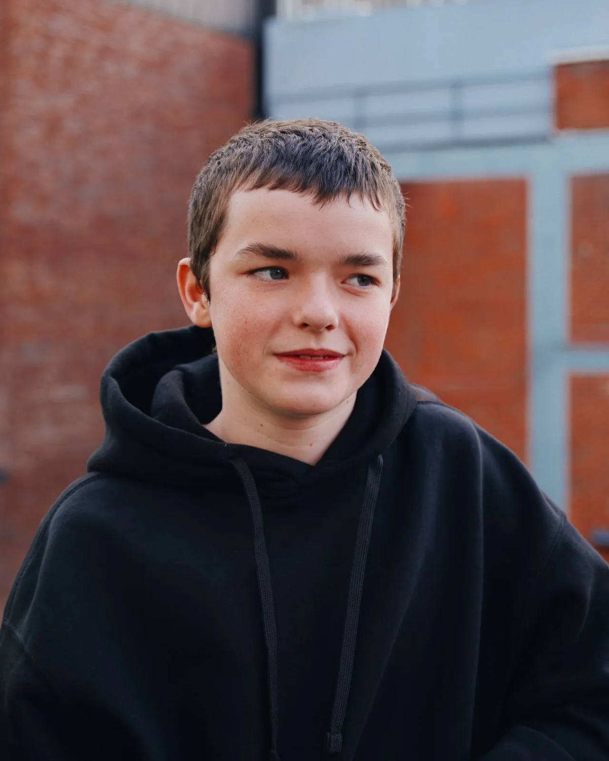 Aktor Owen Cooper z serialu Netflix „Dojrzewanie” w czarnej bluzie z kapturem, uśmiechający się na tle ceglanej ściany.