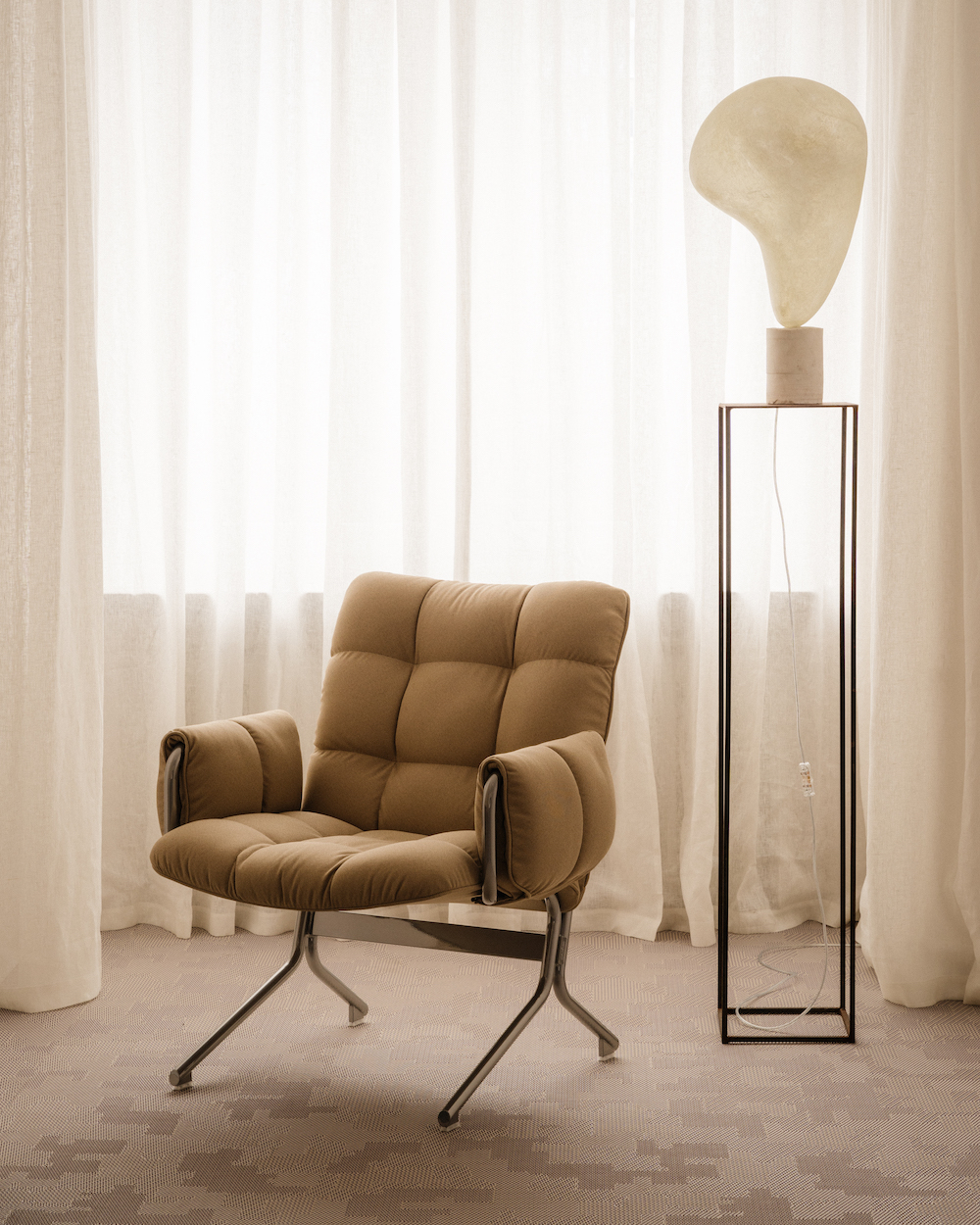 Wnętrze z beżowym fotelem i nowoczesną lampą na tle jasnych firanek. Stonowana kolorystyka i elegancki design.