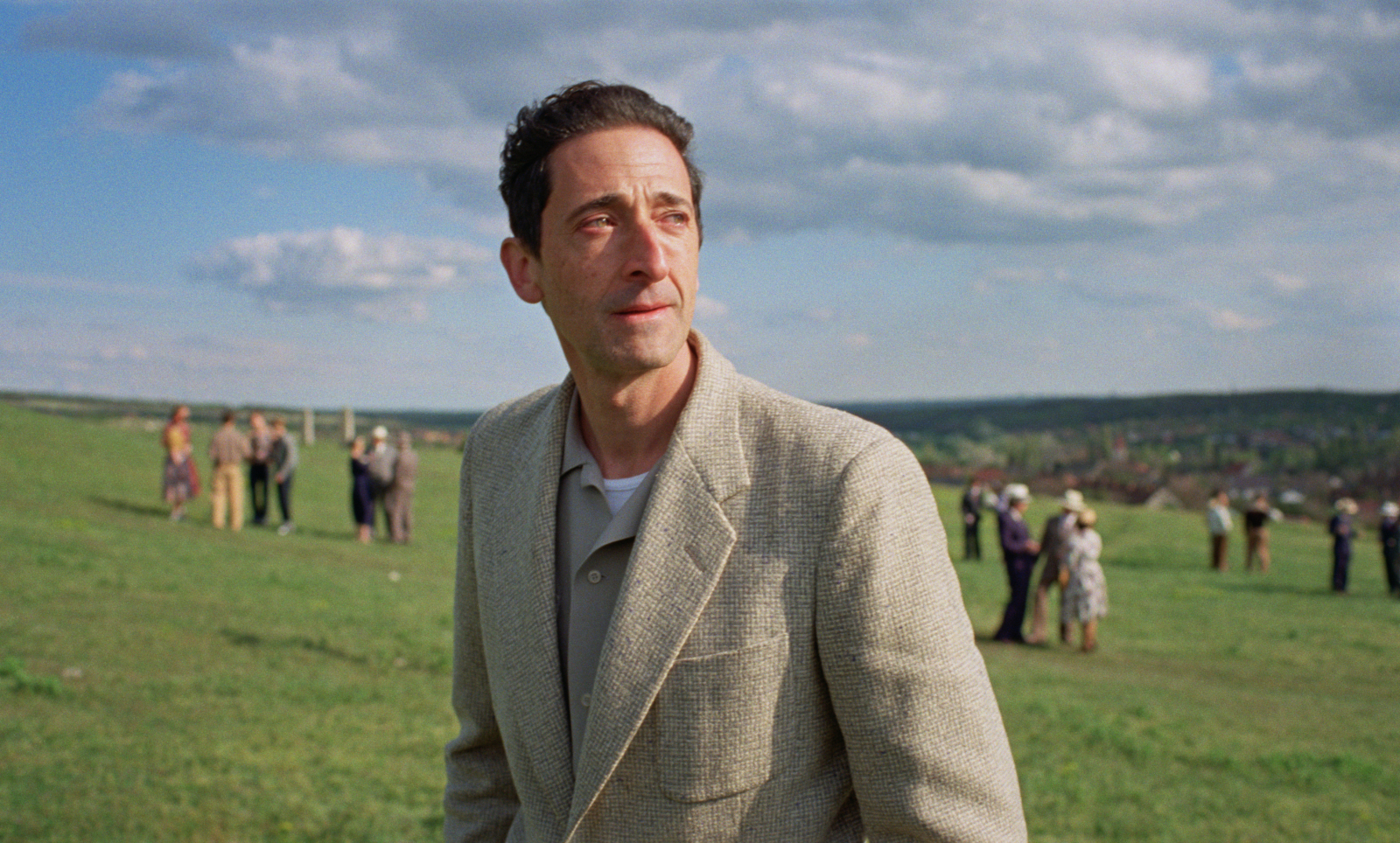 Adrien Brody w scenerii filmu „The Brutalist”, stojący na zielonym wzgórzu w otoczeniu ludzi.
