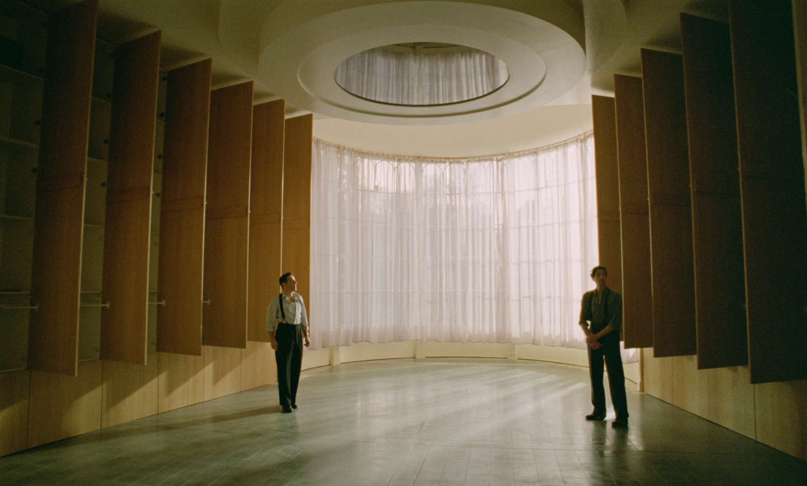 Guy Pearce i Adrien Brody w nowoczesnym wnętrzu w filmie „The Brutalist”, z dużymi oknami i minimalistycznym designem.