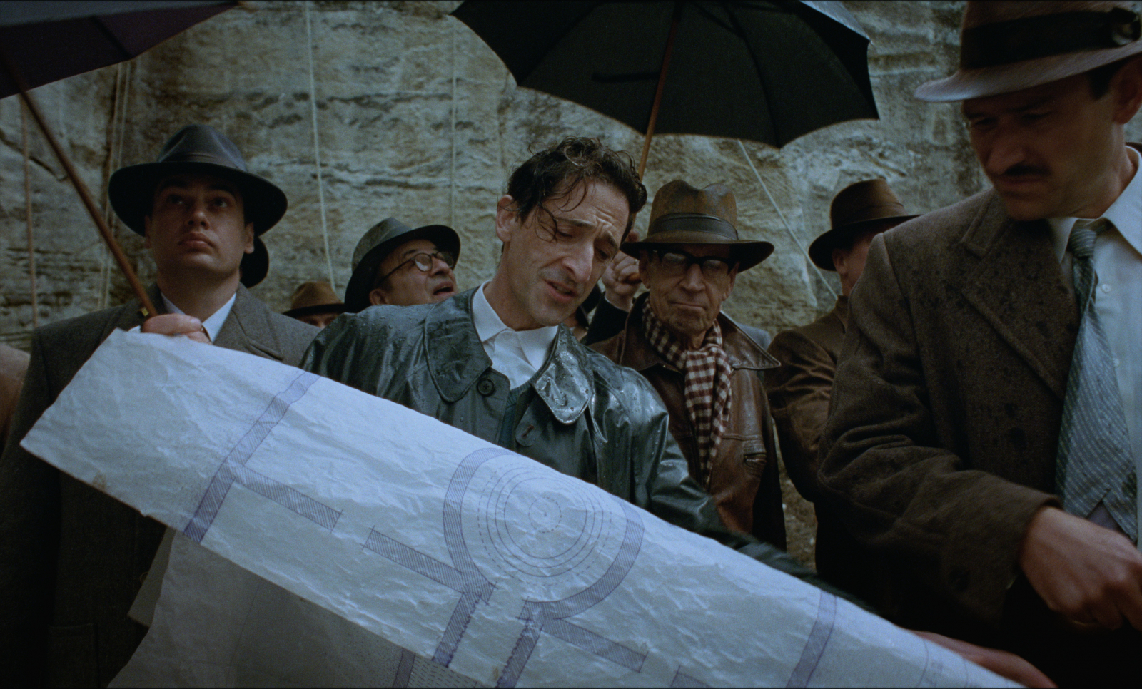 Adrien Brody w filmie „The Brutalist”, trzymając plany budowy, otoczony przez mężczyzn w kapeluszach, w deszczowej scenerii.