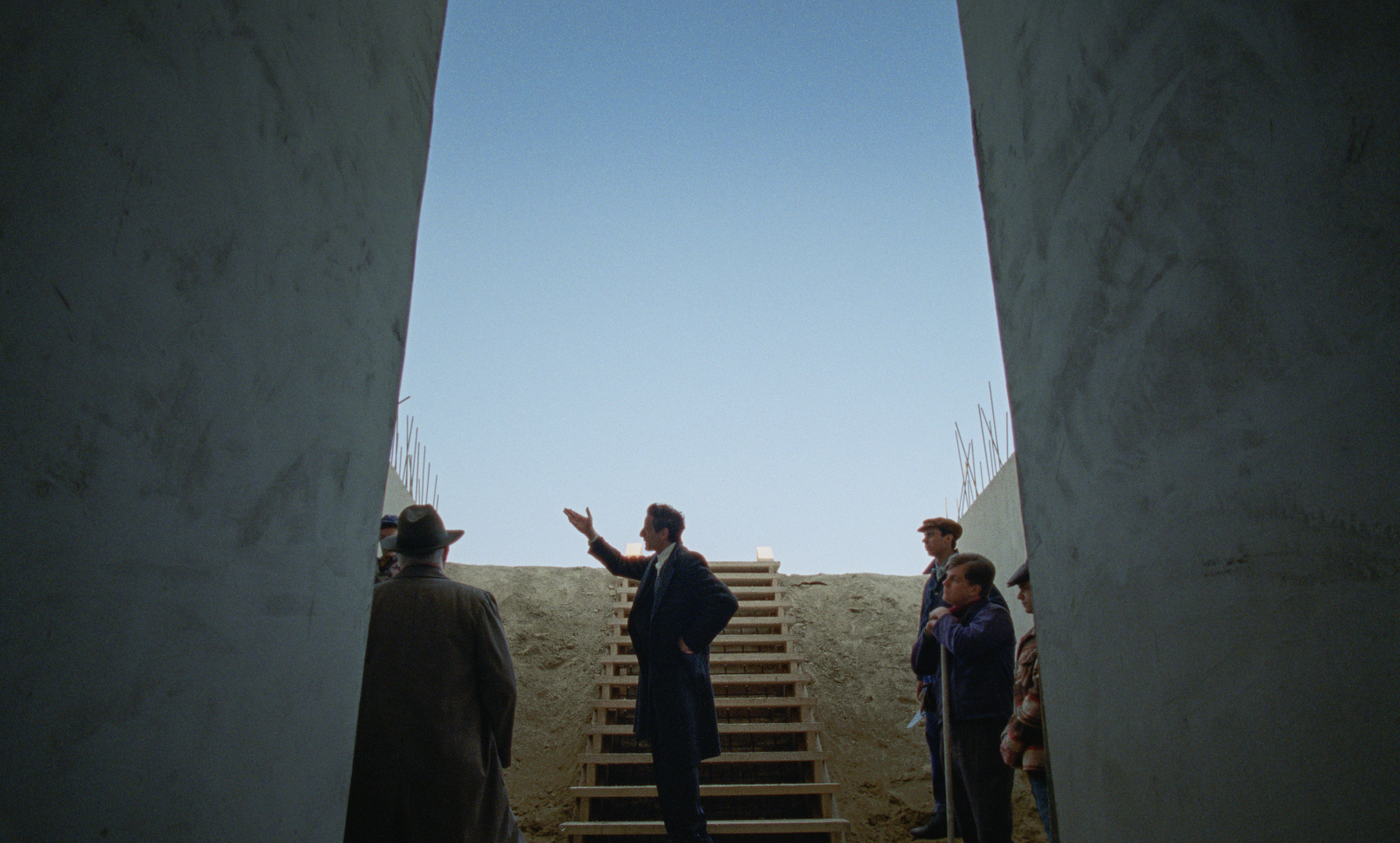 Adrien Brody prowadzi dyskusję w scenie z filmu „The Brutalist” przed betonową konstrukcją i schodami.