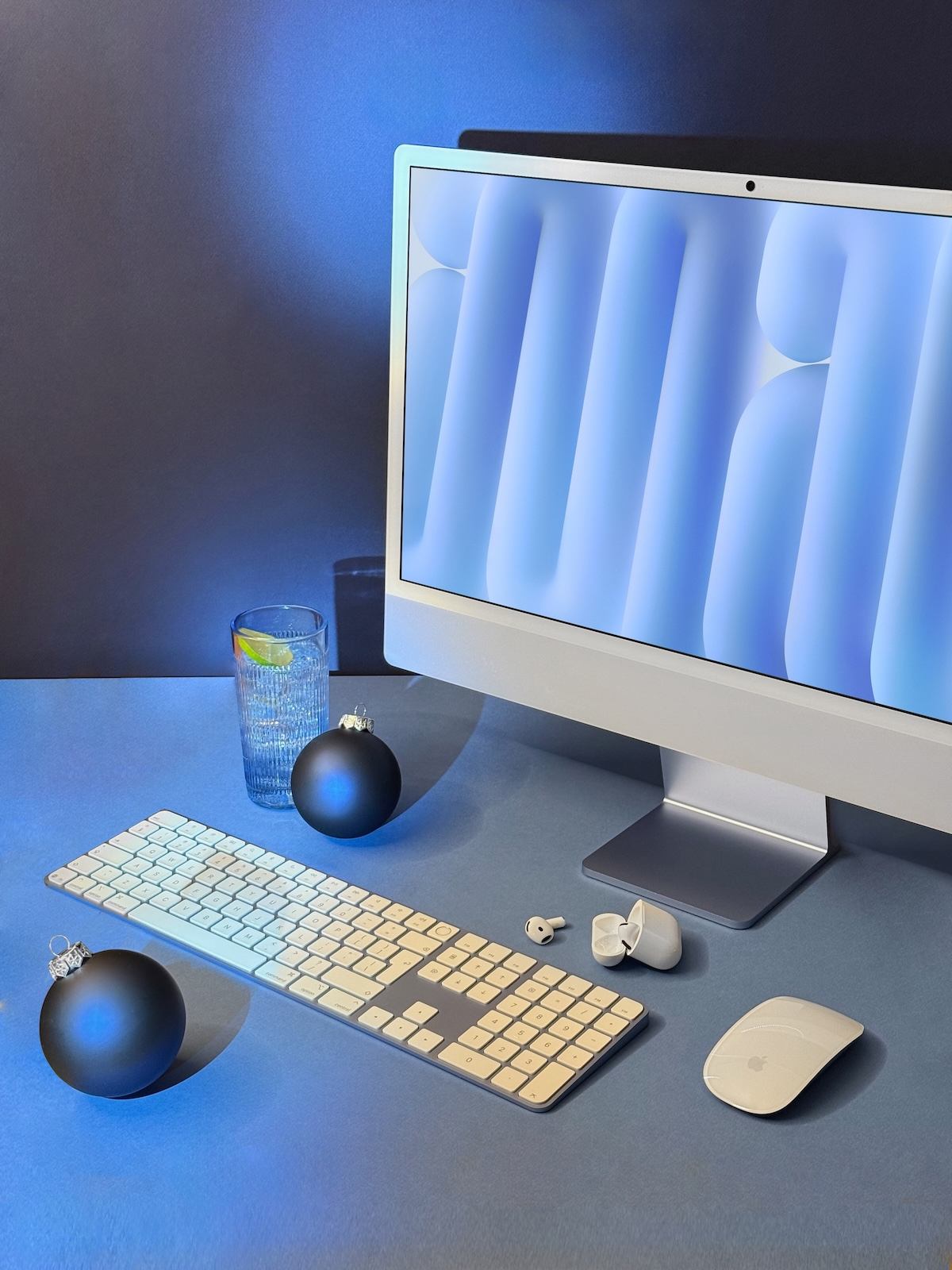 Komputer iMac M4, klawiatura i myszka, podświetlone błękitnym światłem, w otoczeniu błękitnych bombek choinkowych, obok szklanka wody.