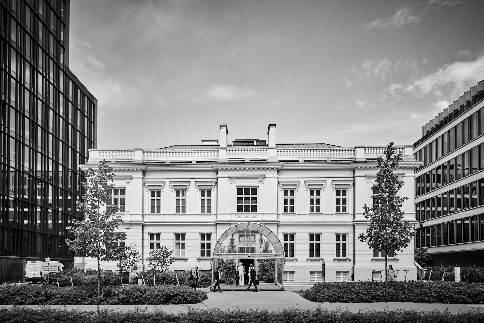 Budynek Archicom: budowla z białą elewacją pośrodku nowoczesnych biurowców, otoczona zielenią.