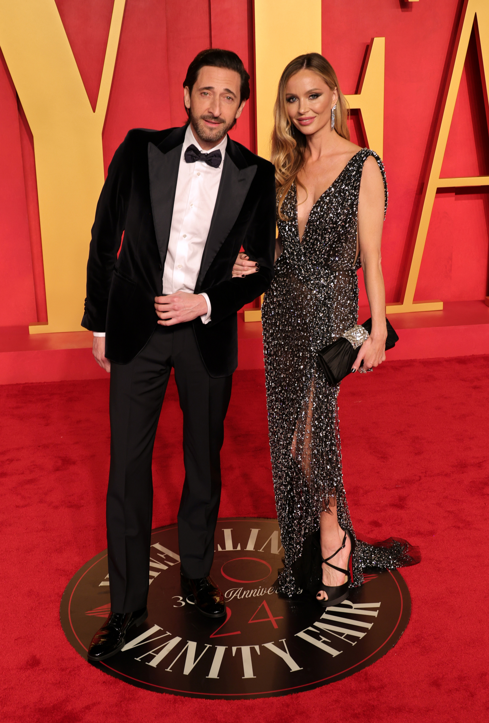 Adrien Brody i Georgina Chapman pozują na czerwonym dywanie, ubrani w eleganckie stroje, przy dużym znaku Vanity Fair.