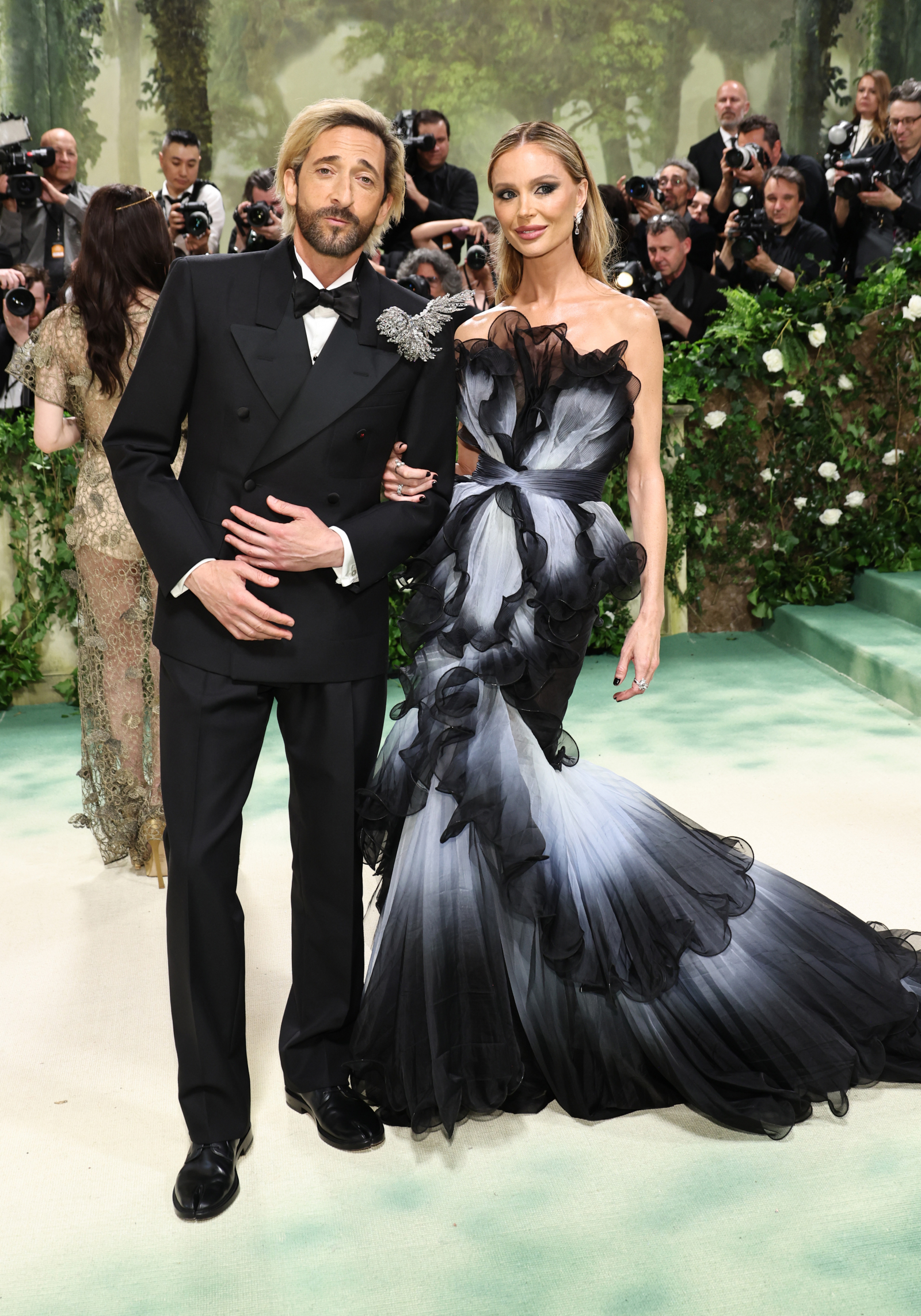 Adrien Brody i Georgina Chapman na Met Gala 2024, w eleganckich strojach na tle zielonej scenerii.