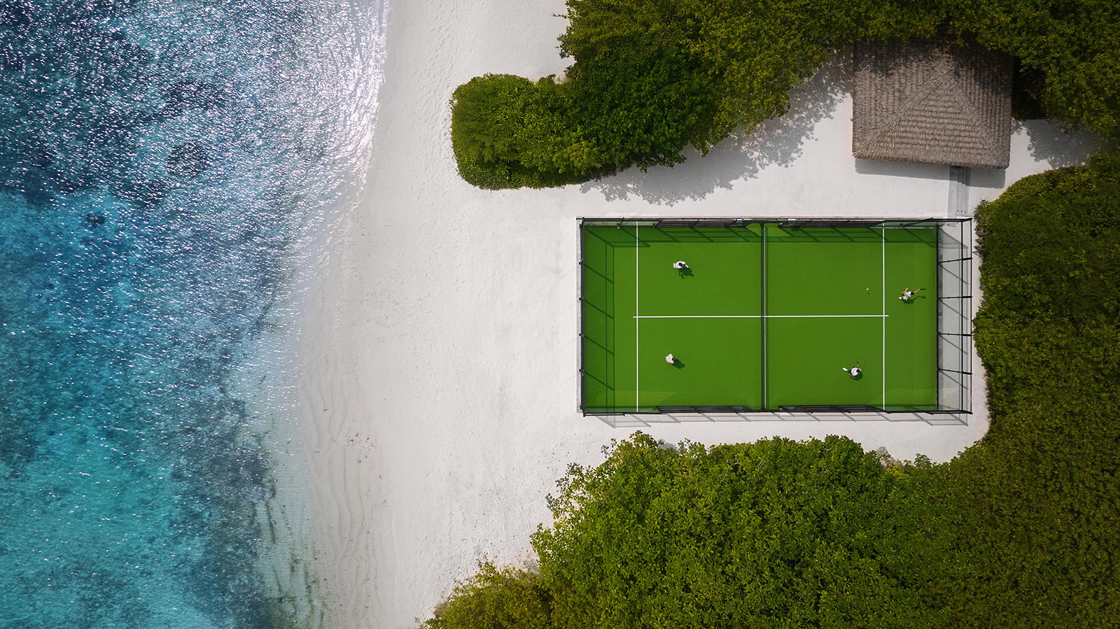 Teren sportowy z zielonym kortem tenisowym otoczony plażą i turkusową wodą. W tle zieleń roślinności i drewniana altana.