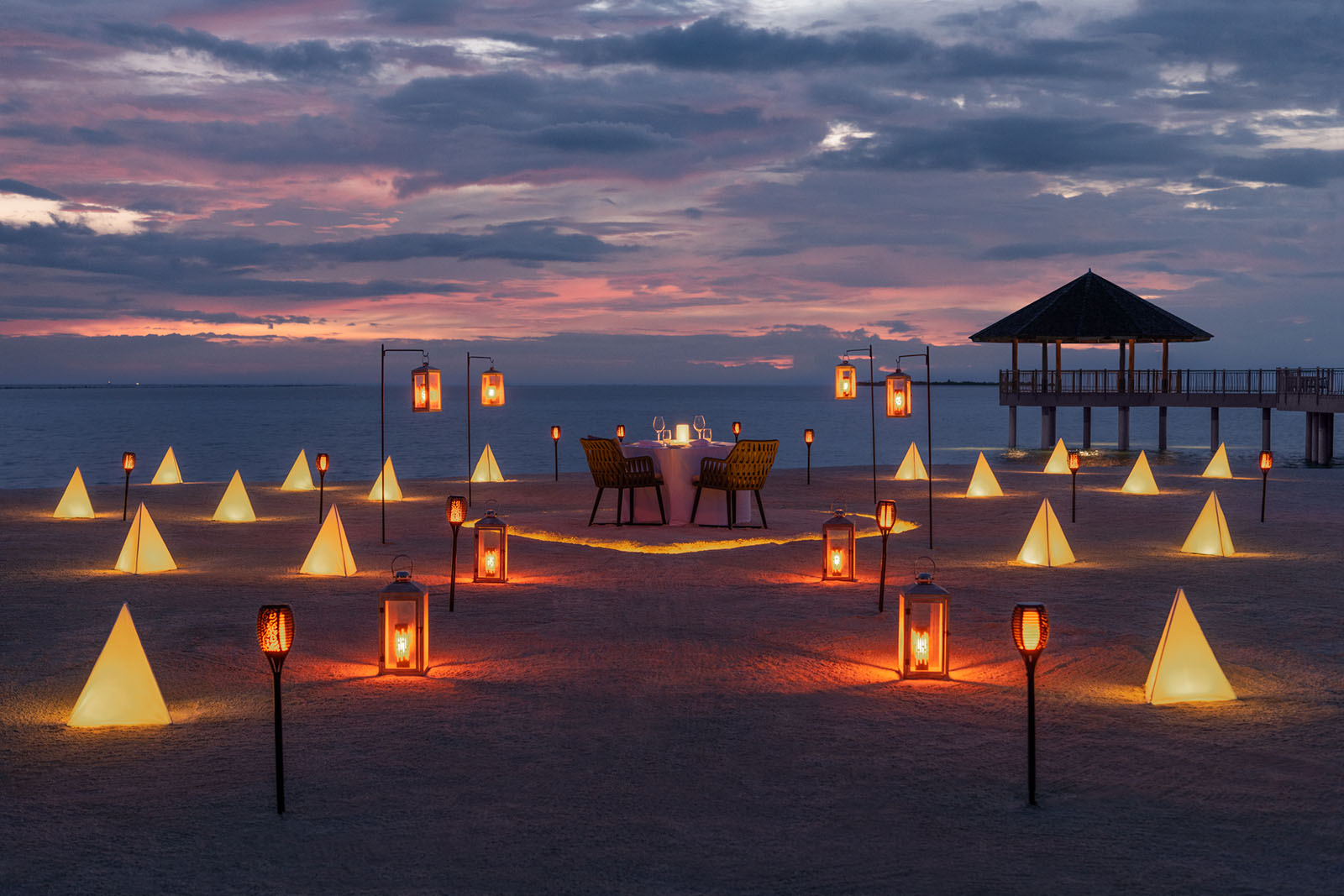 Romantyczna kolacja na plaży przy zachodzie słońca, otoczona podświetlonymi geometrycznymi kształtami i lampionami.