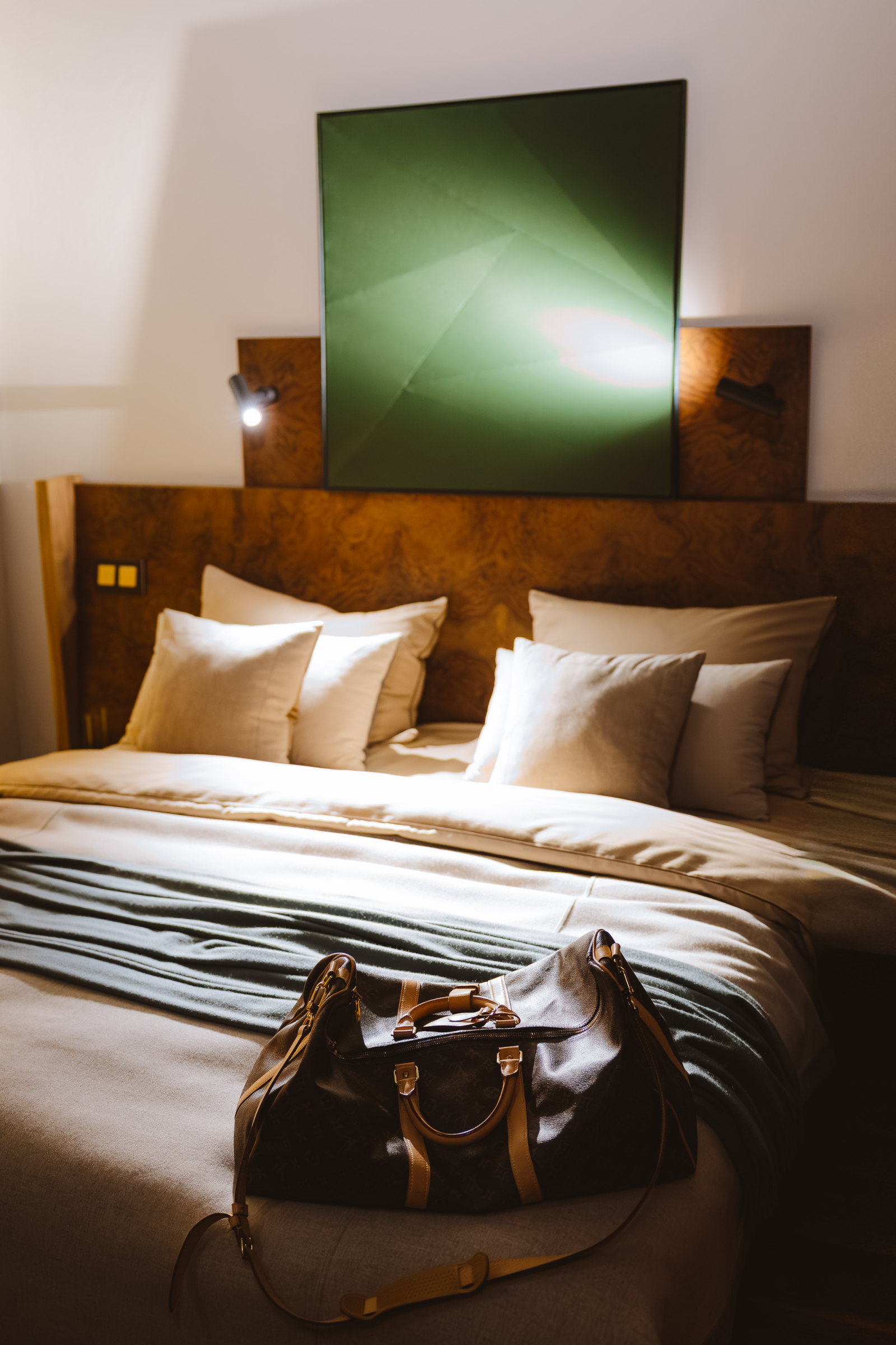 Wnętrze pokoju w Hotelu Warszawa, z dużym łóżkiem, poduszkami i torbą podróżną na pościeli. Na ścianie zielony obraz.