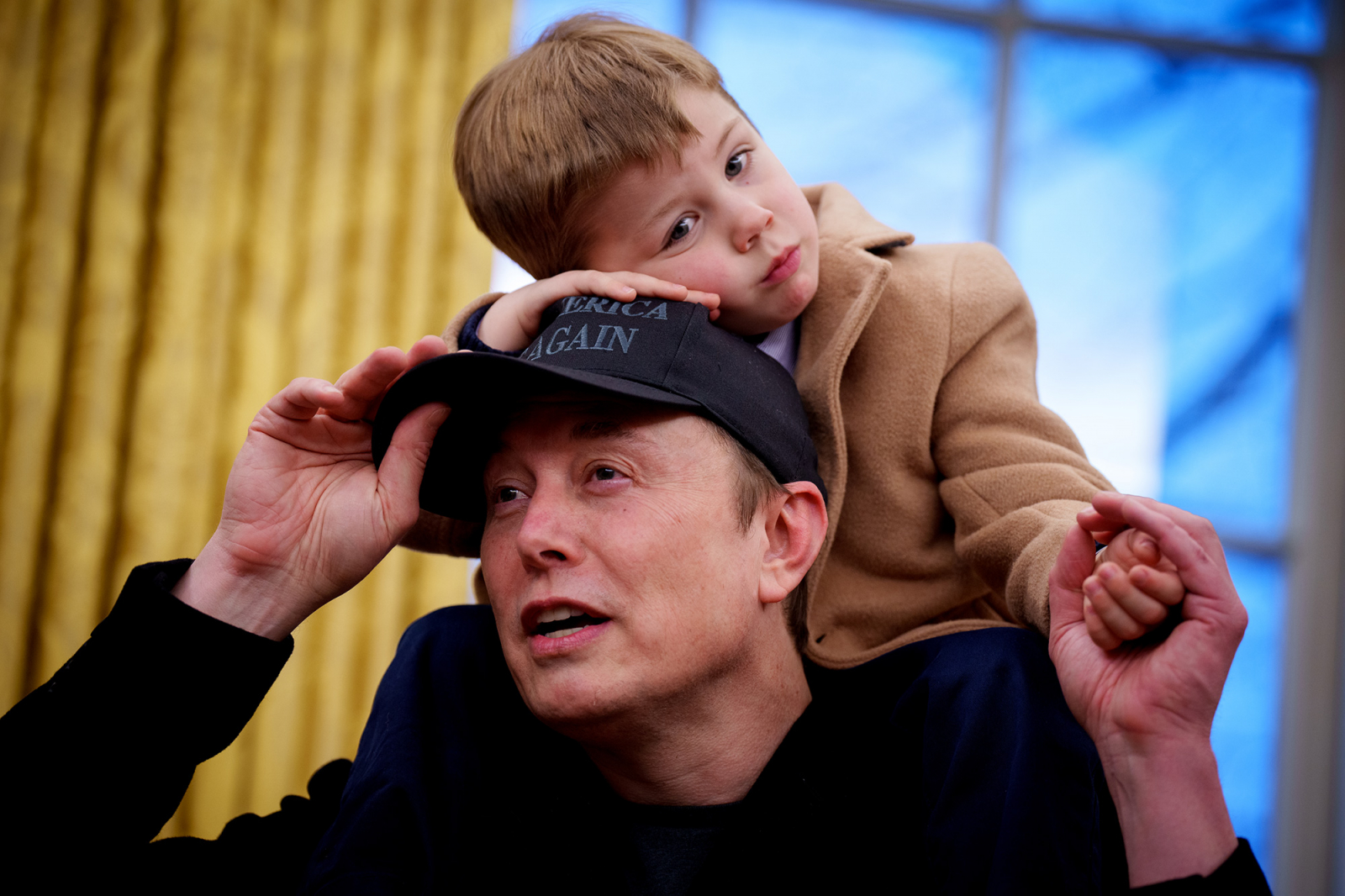 Elon Musk z synem Xem na plecach, oboje uśmiechają się. Tło to zasłony w odcieniach złota. Dziecko nosi brązowy płaszcz.