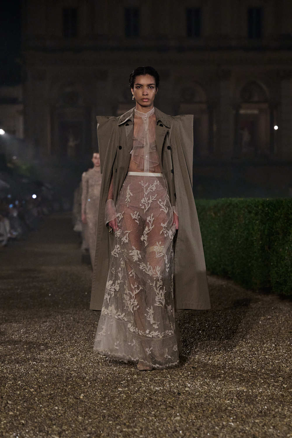 Modelka w koronkowej sukience na lato, z przezroczystymi elementami, idzie w eleganckiej scenerii na pokazie ślubnym.