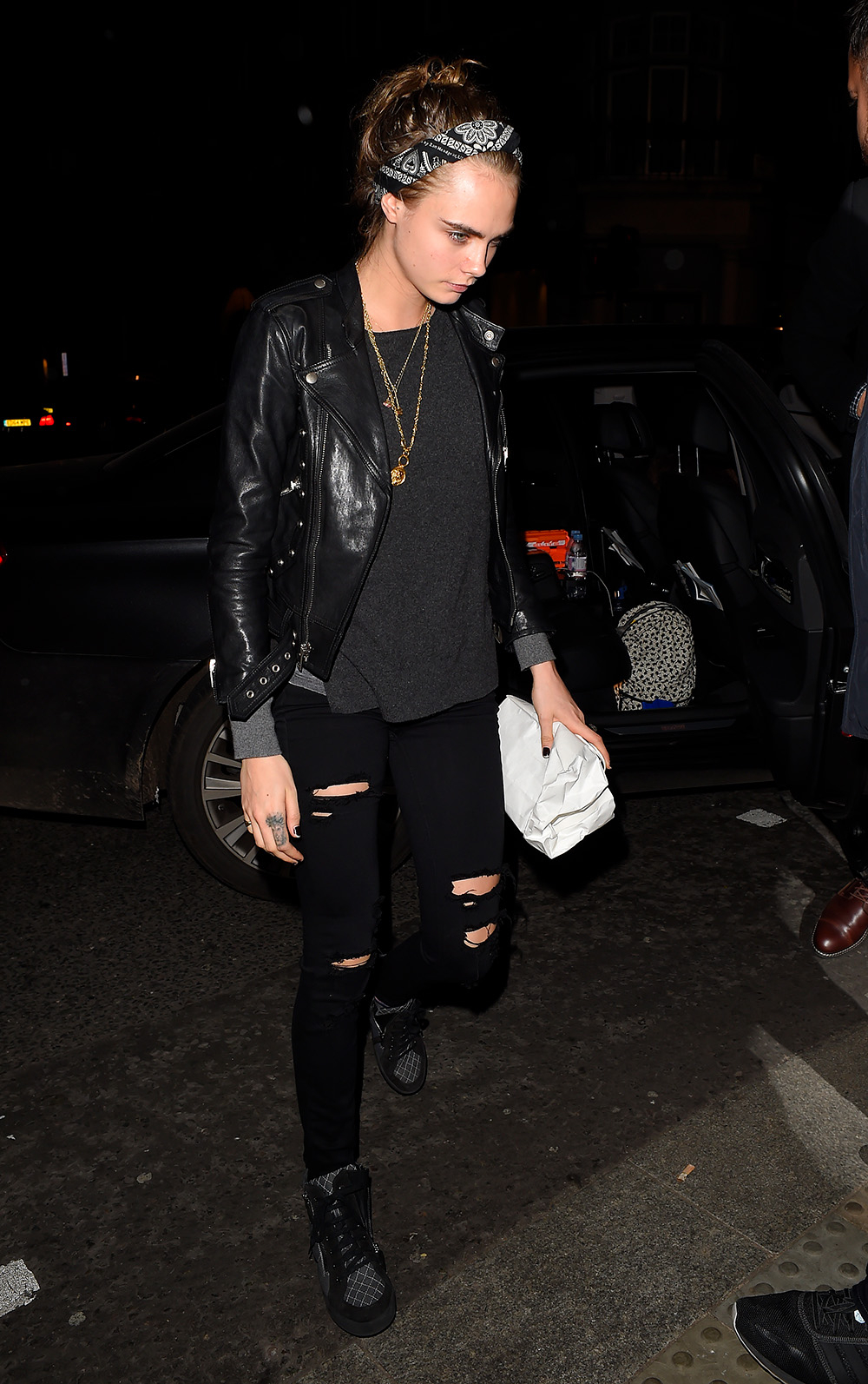 Cara Delevingne w czarnej skórzanej kurtce i bandance, trzyma torebkę, idąc w nocy w mieście.