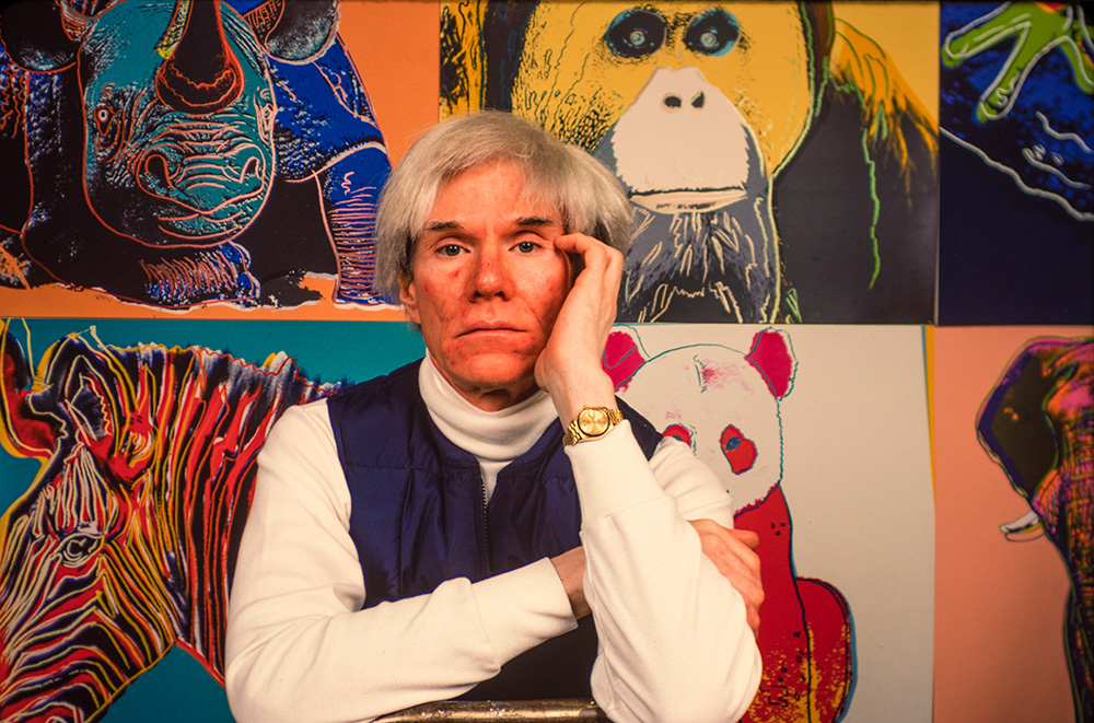 Andy Warhol pozując wśród kolorowych artystycznych prac przedstawiających zwierzęta, z ręką na twarzy.