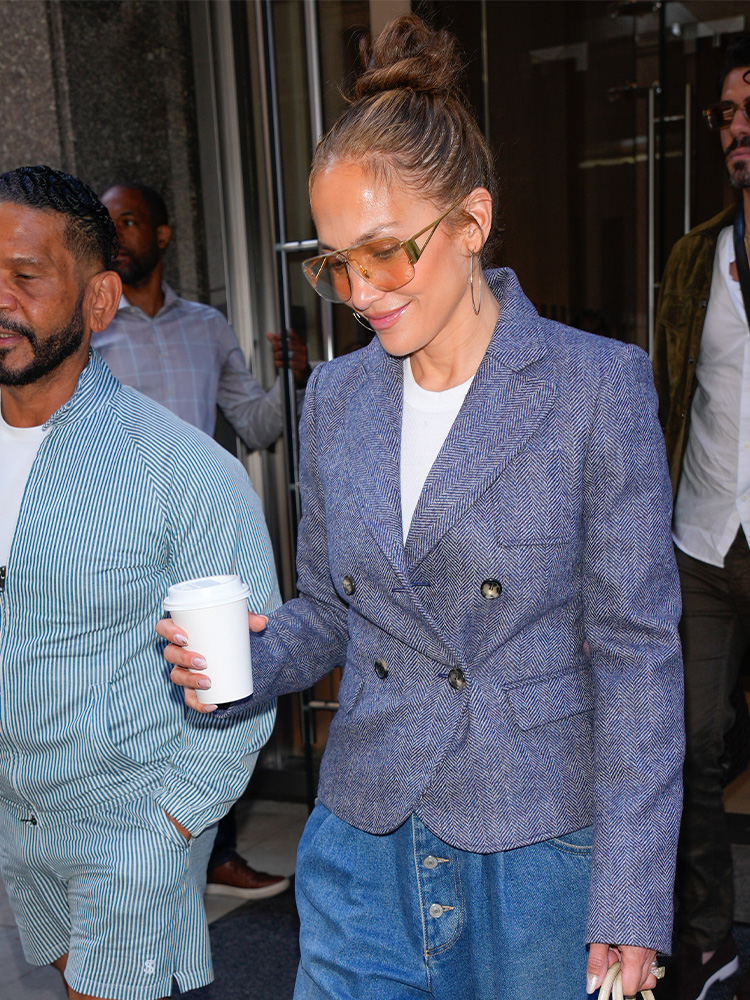 Jennifer Lopez w eleganckim żakiecie i baggy jeans, trzyma filiżankę kawy, uśmiechając się.