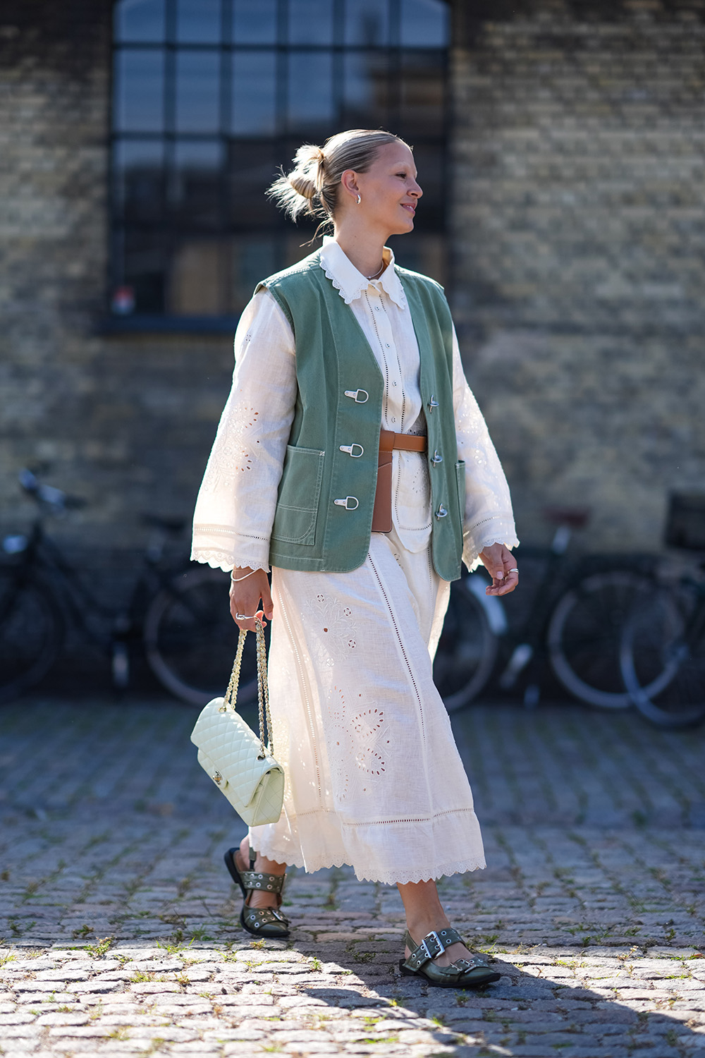 Kobieta w białej długiej spódnicy oraz zielonej kamizelce, spacerująca z torebką w stylu vintage.