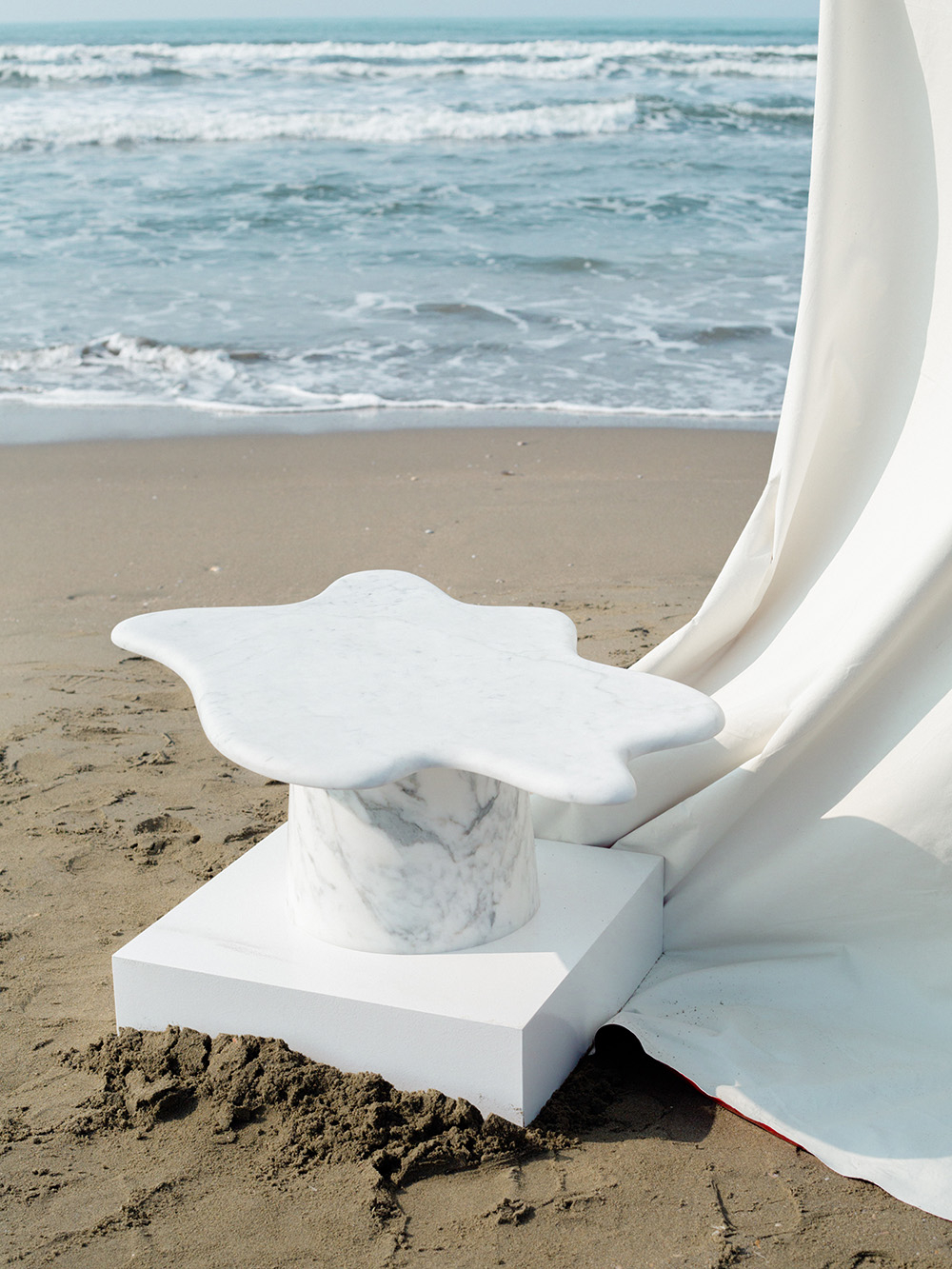 Marble table o nietypowym kształcie, umieszczony na plaży, przy morzu, obok białego materiału.