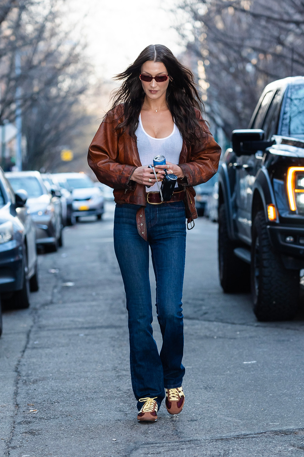 Bella Hadid w brązowych adidasach, w dżinsach i skórzanej kurtce, spaceruje po ulicy w miejskim otoczeniu.