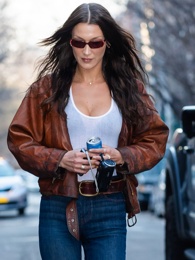Bella Hadid w brązowej skórzanej kurtce i niebieskich dżinsach, trzyma puszkę napoju i aparat na ulicy.