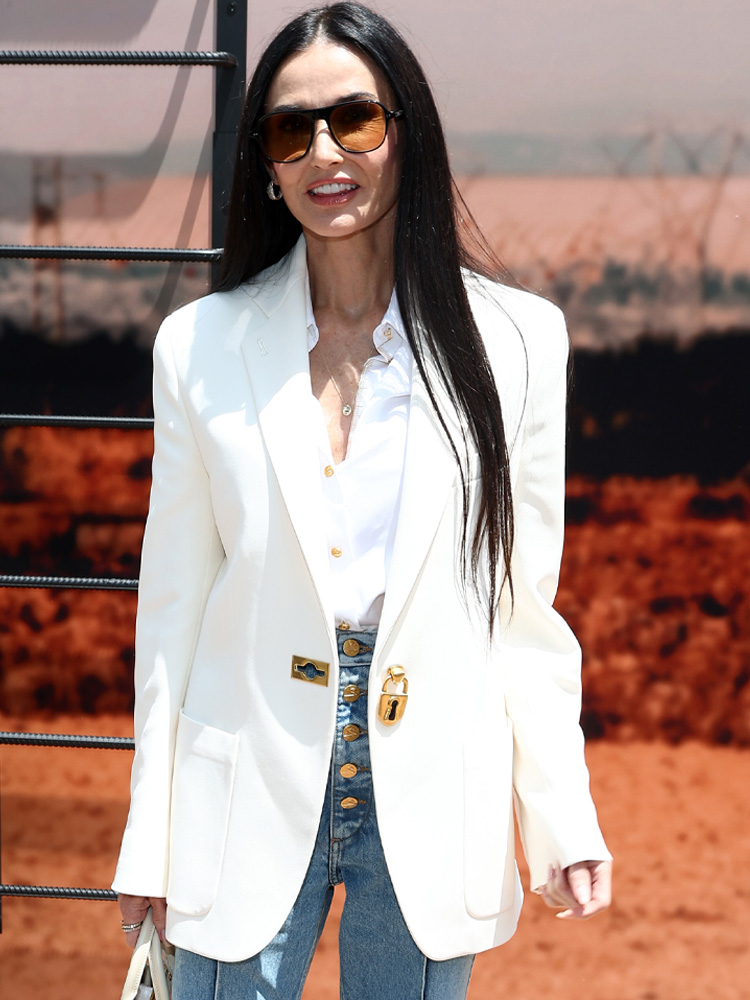 Demi Moore w białej marynarce, białej koszuli i niebieskich dżinsach, uśmiechając się z okularami przeciwsłonecznymi.