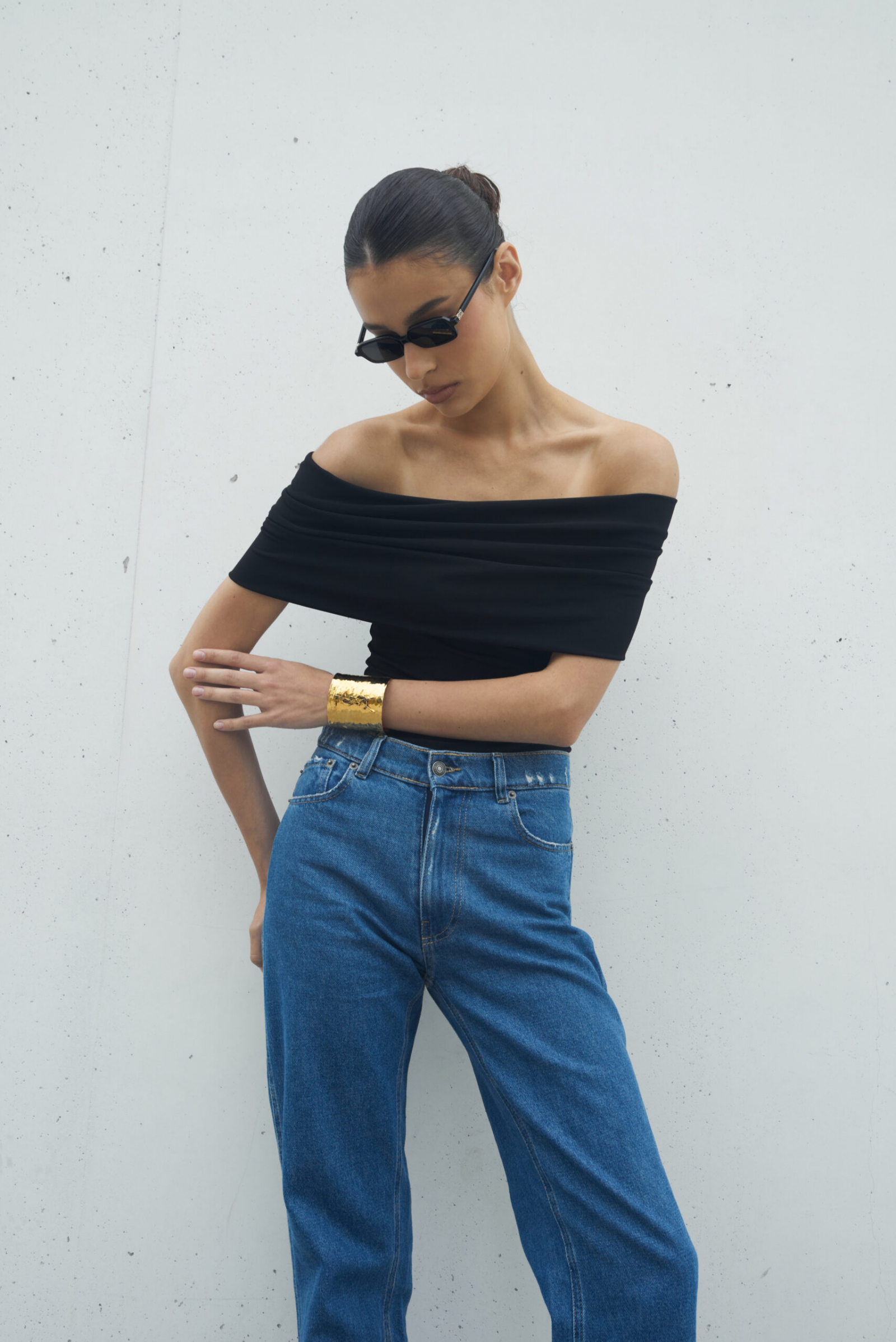 Modelka w czarnej, off-shoulder bluzce i niebieskich dżinsach, wystylizowana z dodatkiem złotej bransolety, pozująca na tle ściany.