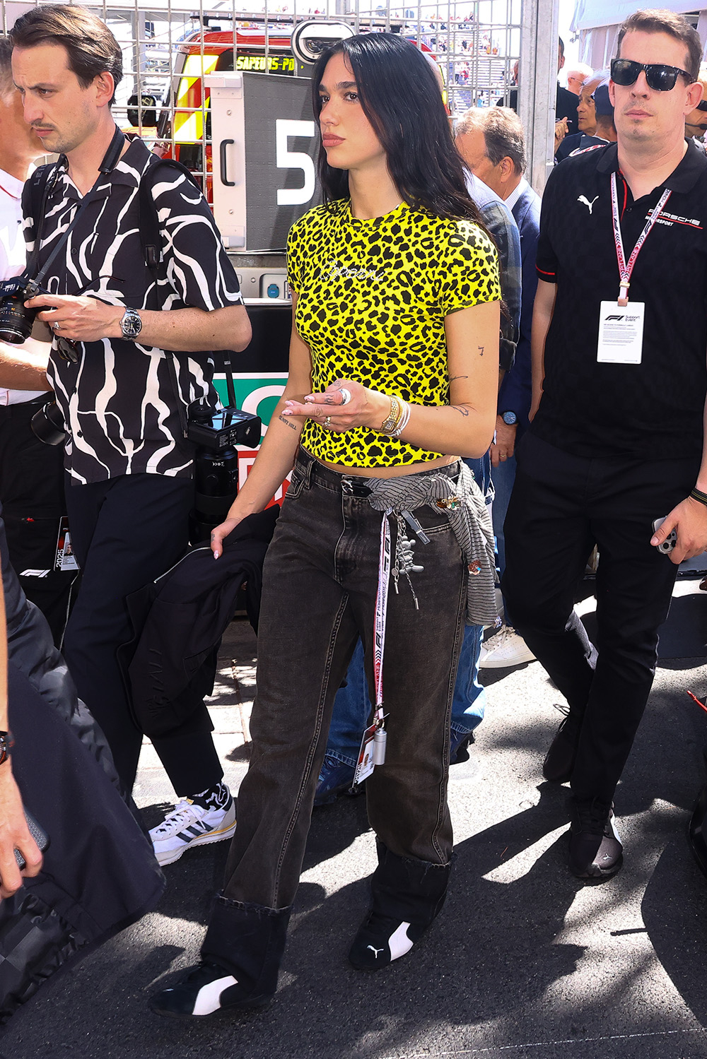 Dua Lipa w jaskrawym, młodzieżowym stroju, przechodzi obok grupy ludzi na wydarzeniu.