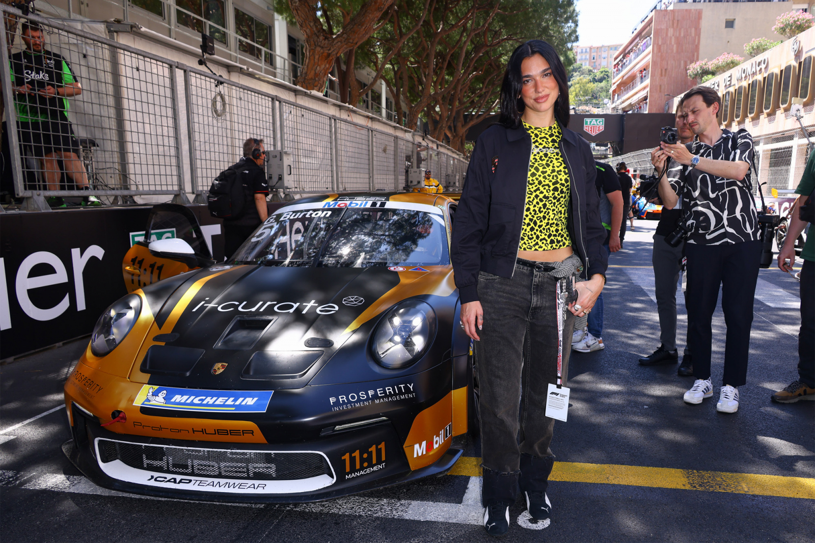 Dua Lipa stoi obok wyścigowego samochodu Porsche na torze, ubrana w modny zestaw. W tle widać innych ludzi.