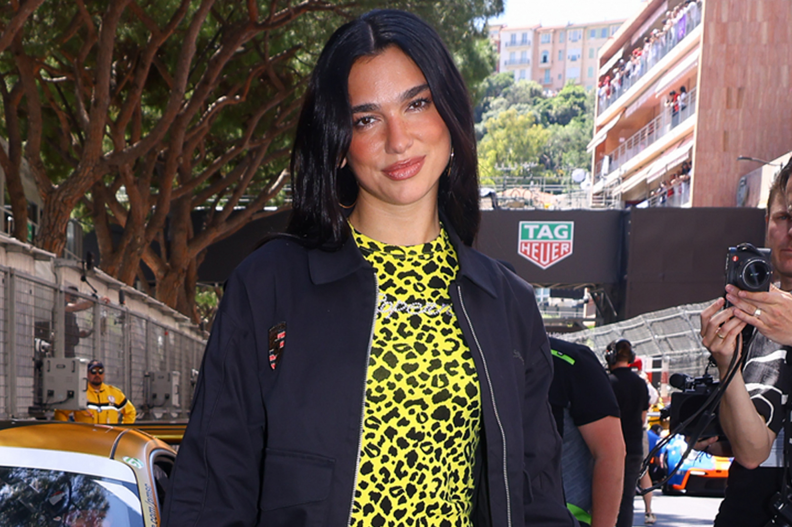 Dua Lipa w kolorowej, wzorzystej koszulce i czarnym płaszczu, na tle wyścigowego toru.