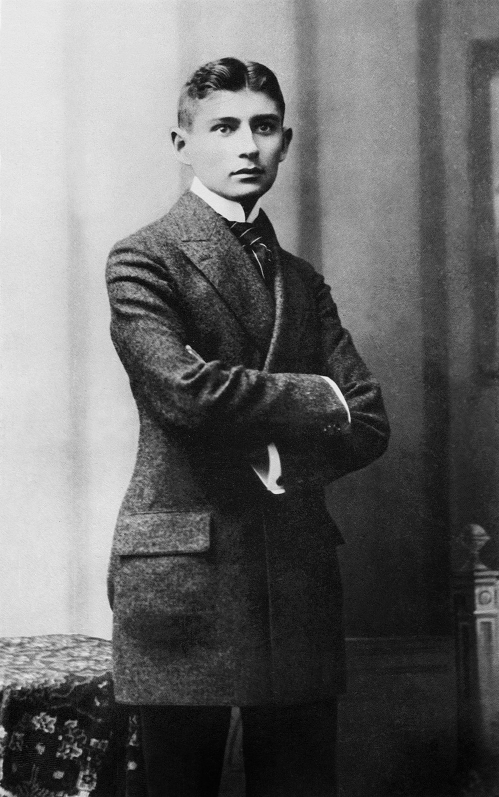 Franz Kafka w młodości, ubrany w elegancki garnitur, stoi z założonymi rękami.