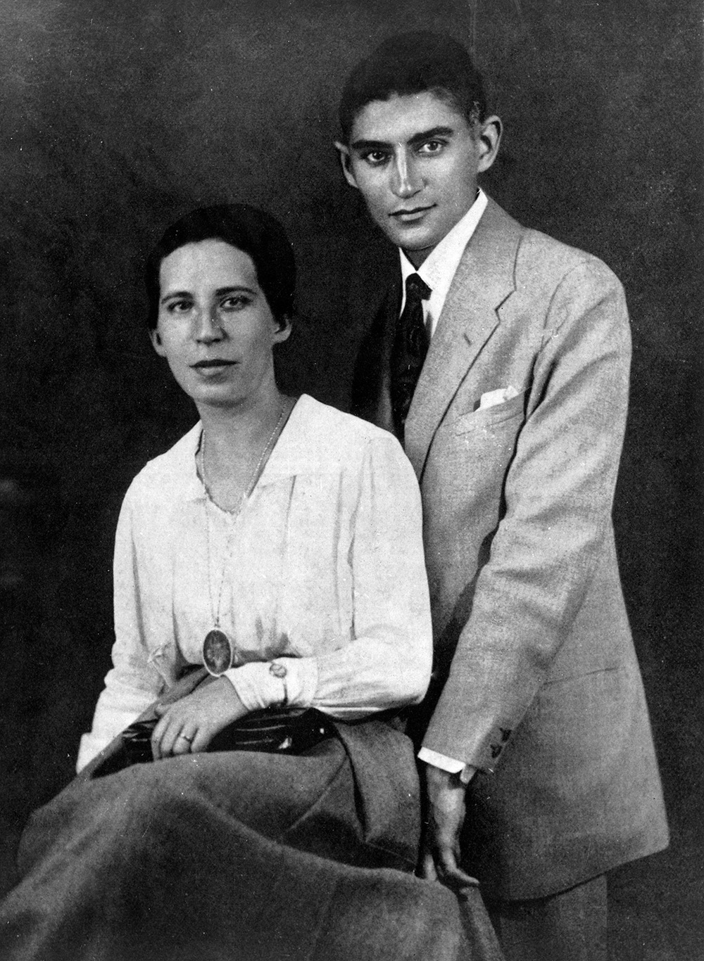 Franz Kafka z narzeczoną w eleganckim stroju, pozujący na tle ciemnej ściany. Ona siedzi, on stoi za nią.