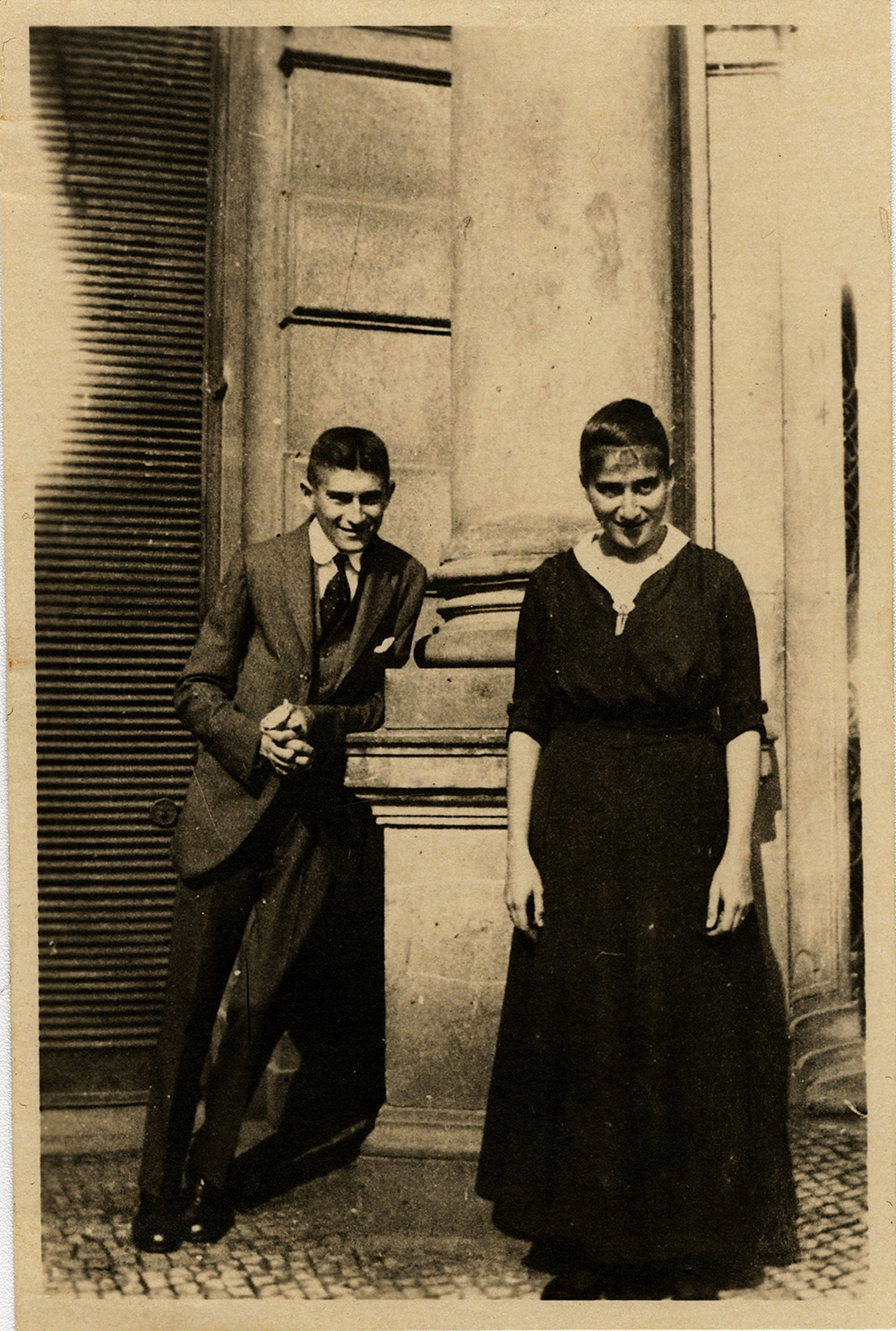 Franz Kafka z siostrą stoją przy kolumnie, uśmiechnięci, w starym budynku. On w eleganckim garniturze, ona w czarnej sukni.