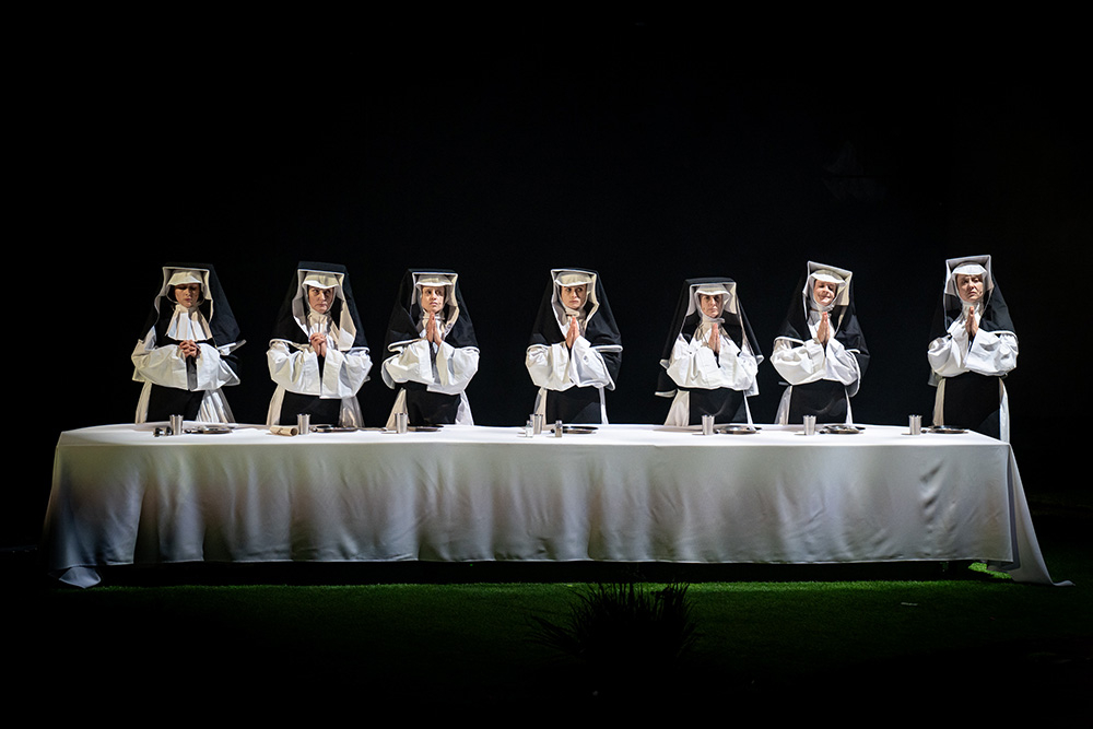 Siedem zakonnic w habitach modli się przy długim stole, w ciemnej scenerii.