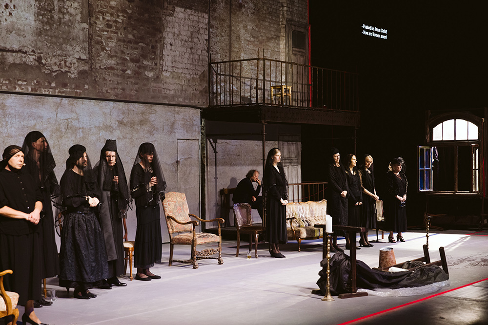 Scena teatralna przedstawiająca grupę postaci w czarnych strojach, stojących na tle minimalistycznej scenografii.