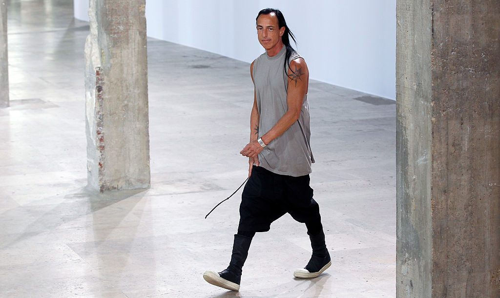 Rick Owens w szarej koszuli bez rękawów i czarnych spodniach, chodzi po minimalistycznym wnętrzu.