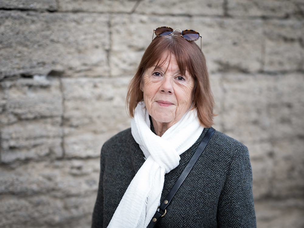 Gisèle Pelicot stoi przy kamiennej ścianie, w ciemnym płaszczu i białym szaliku, w okularach przeciwsłonecznych.
