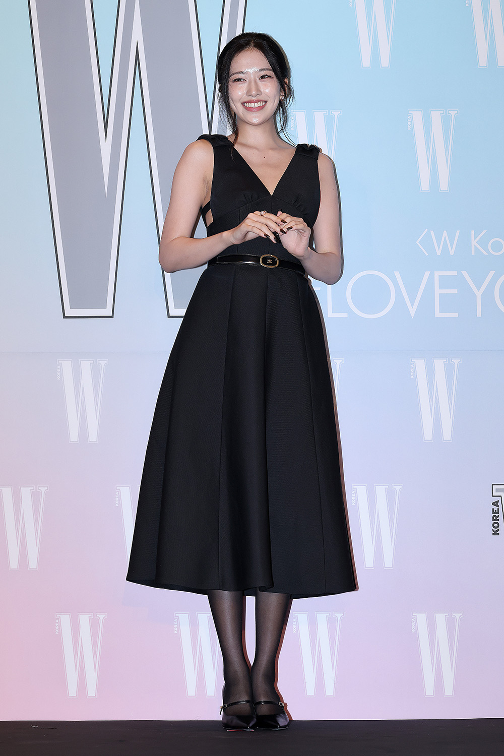 Uśmiechnięta kobieta w czarnej sukience bez rękawów, stojąca na tle pastelowego napisu „W” podczas eventu.