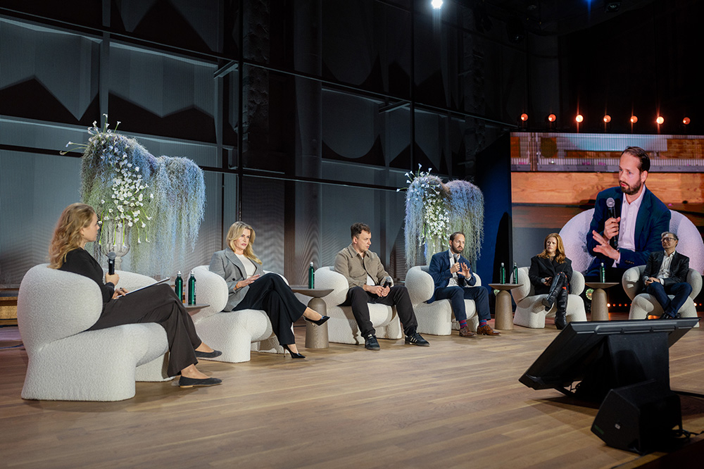 Na scenie siedzi pięciu uczestników panelu dyskusyjnego, z elementami dekoracyjnymi w tle. Na ekranie wyświetlany jest także rozmówca.