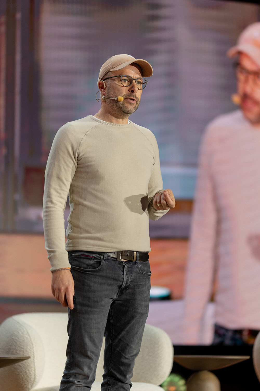 Matthias Haase przemawia na scenie, ubrany w jasny sweter i czapkę, z mikrofonem na głowie.