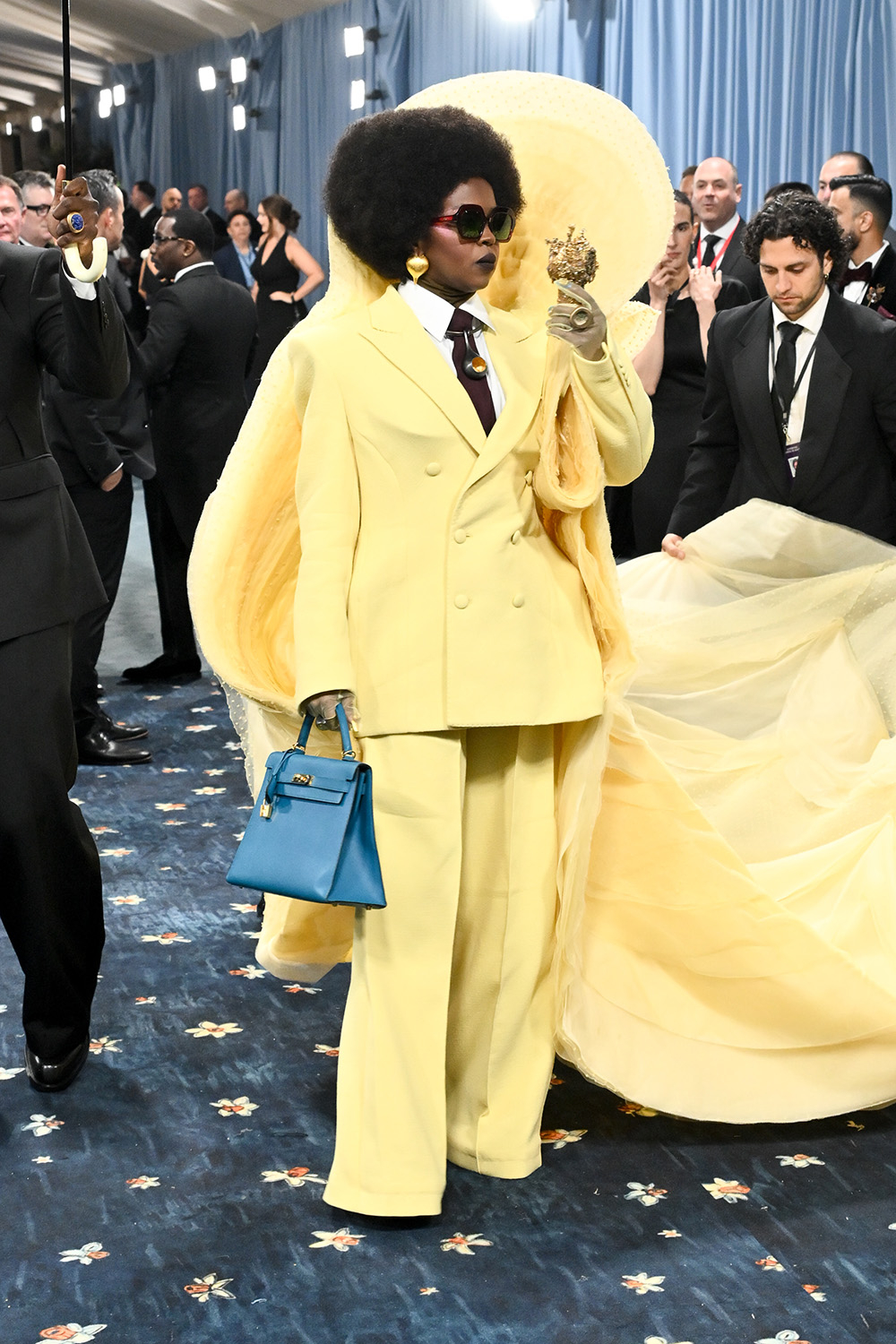 Lauryn Hill w żółtym garniturze z ikonową Birkin bag, w otoczeniu eleganckich gości na wydarzeniu.