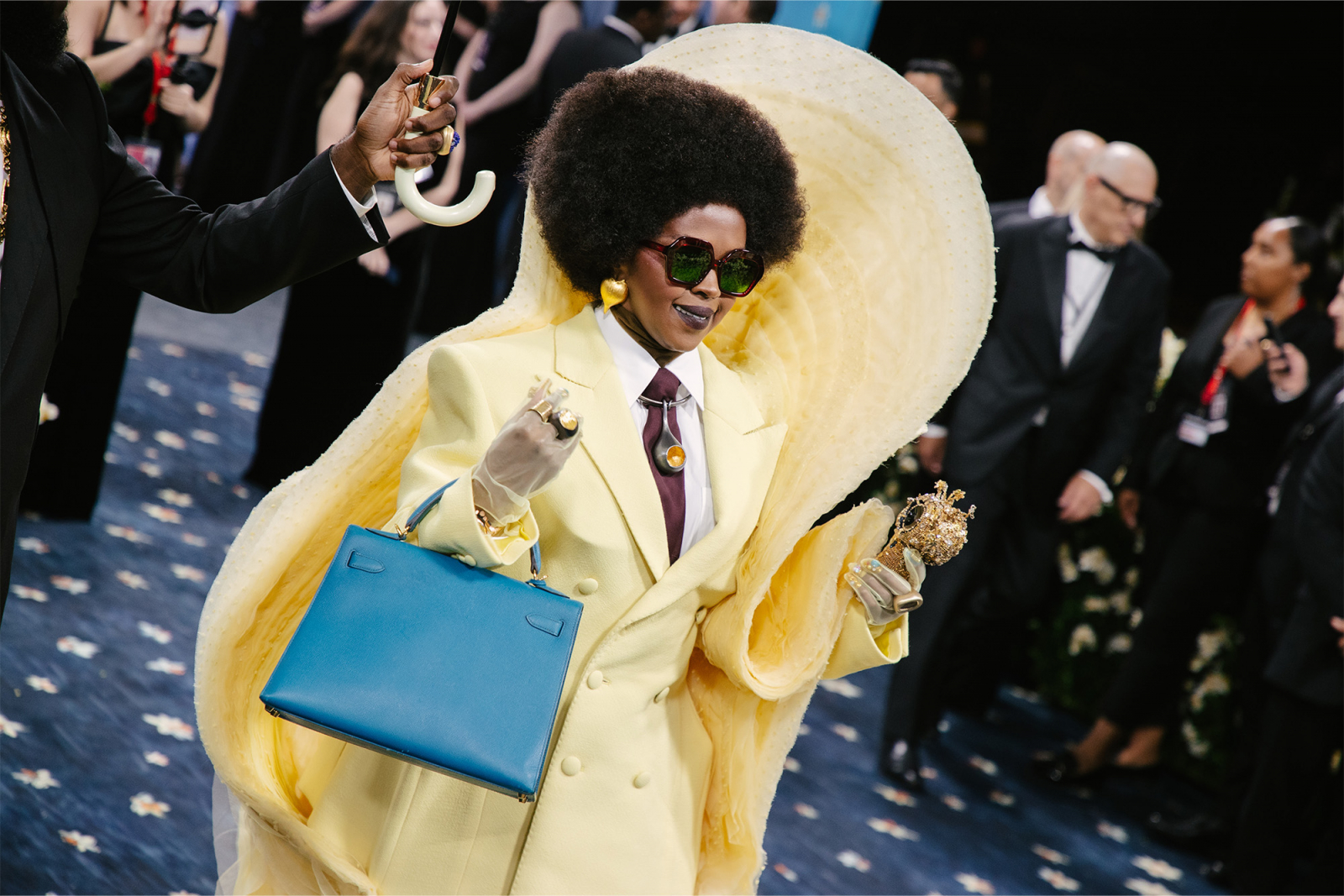 Lauryn Hill w żółtym garniturze i kultową Birkin bag, pod parasolem, pewnie kroczy na czerwonym dywanie.